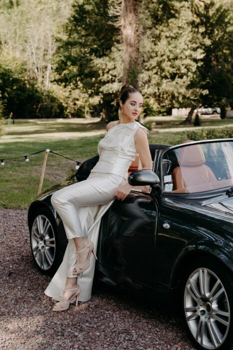Pantalon de la mariée avec un top asymétrique en soie sauvage assise sur la portière d'une voiture